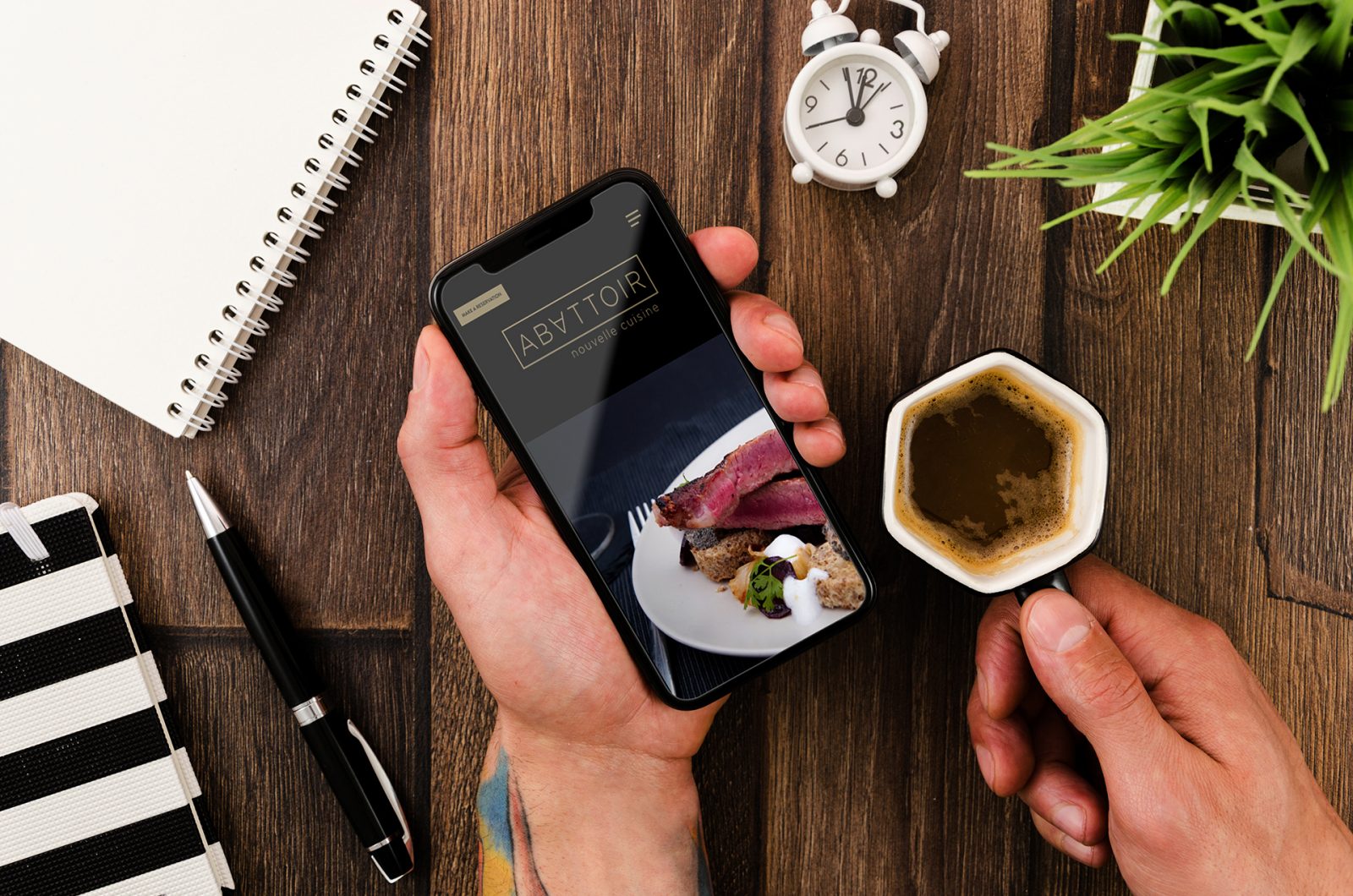 This image shows someone holding a mobile phone with the home page for Abattoir Nouvelle Cuisine. The home page features a black header with a gold wordmark logo as well as a picture of a venison entree.