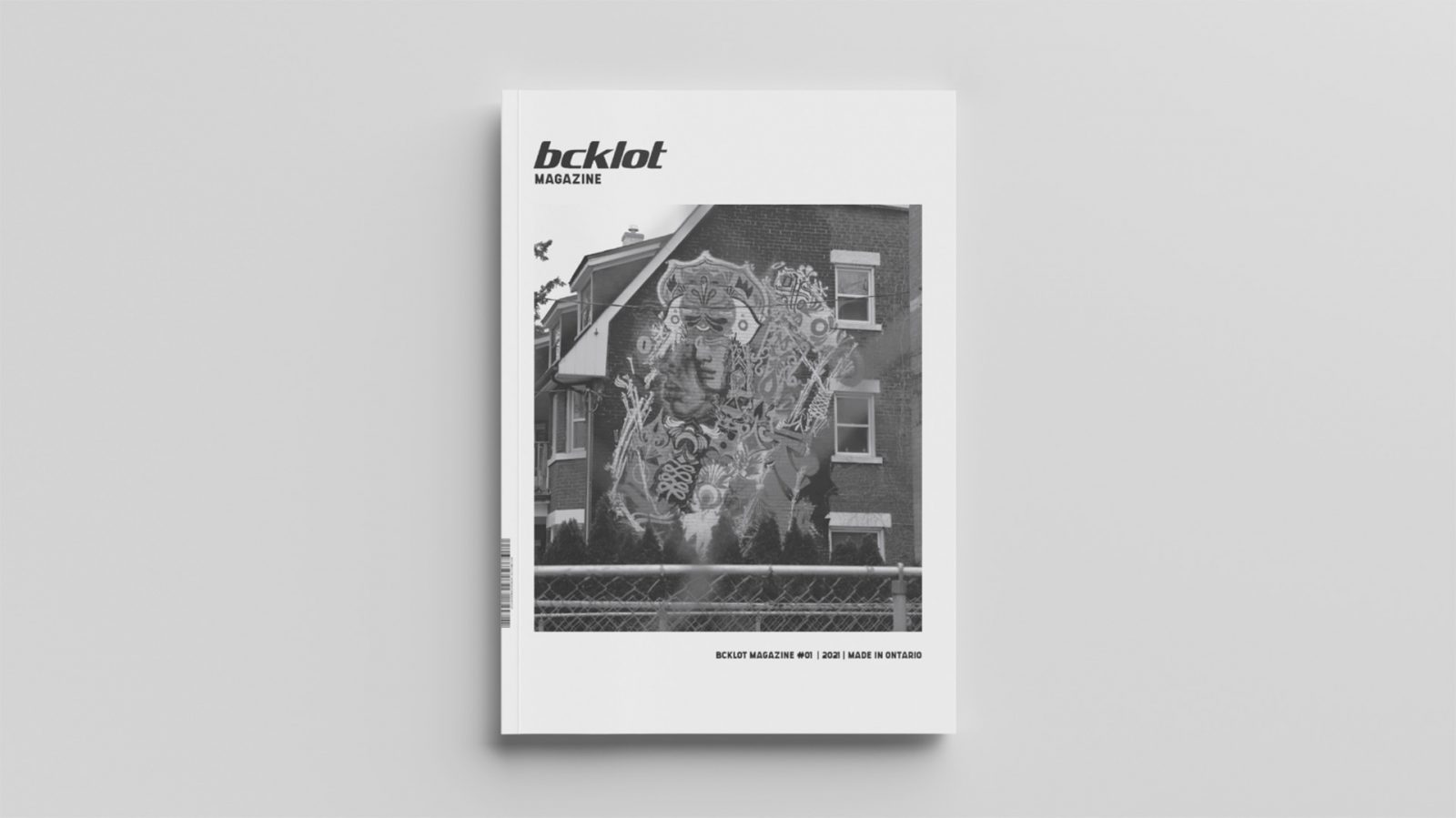 Bcklot (Back-lot) magazine cover. Magazine cover includes centered black and white image of graffiti on the side of home. Bordered in a thick white margin with “Bcklot” logo at top left above image and issue number on bottom right below image. The graffiti on the side of the house illustrates two persons of native heritage and culture wearing headdresses accompanied by cultural symbols and paintings. Image is shot through a wired fence which is visible in the foreground of the image, however out of focus.