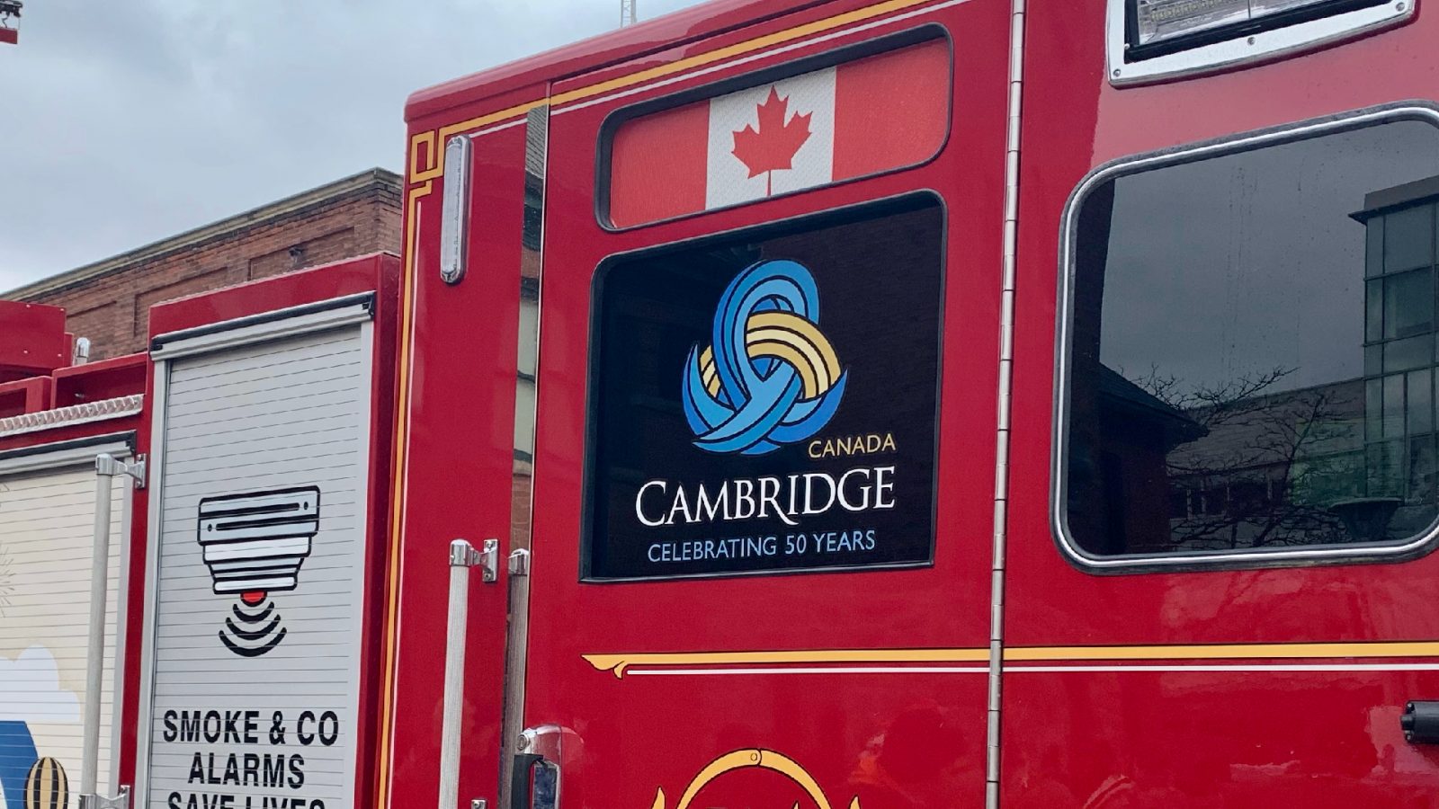 City of Cambridge 50th anniversary logo in vertical format features a blue intertwining loop with a yellow rainbow shape through the loop. It is seen on the side window of a Cambridge fire truck.