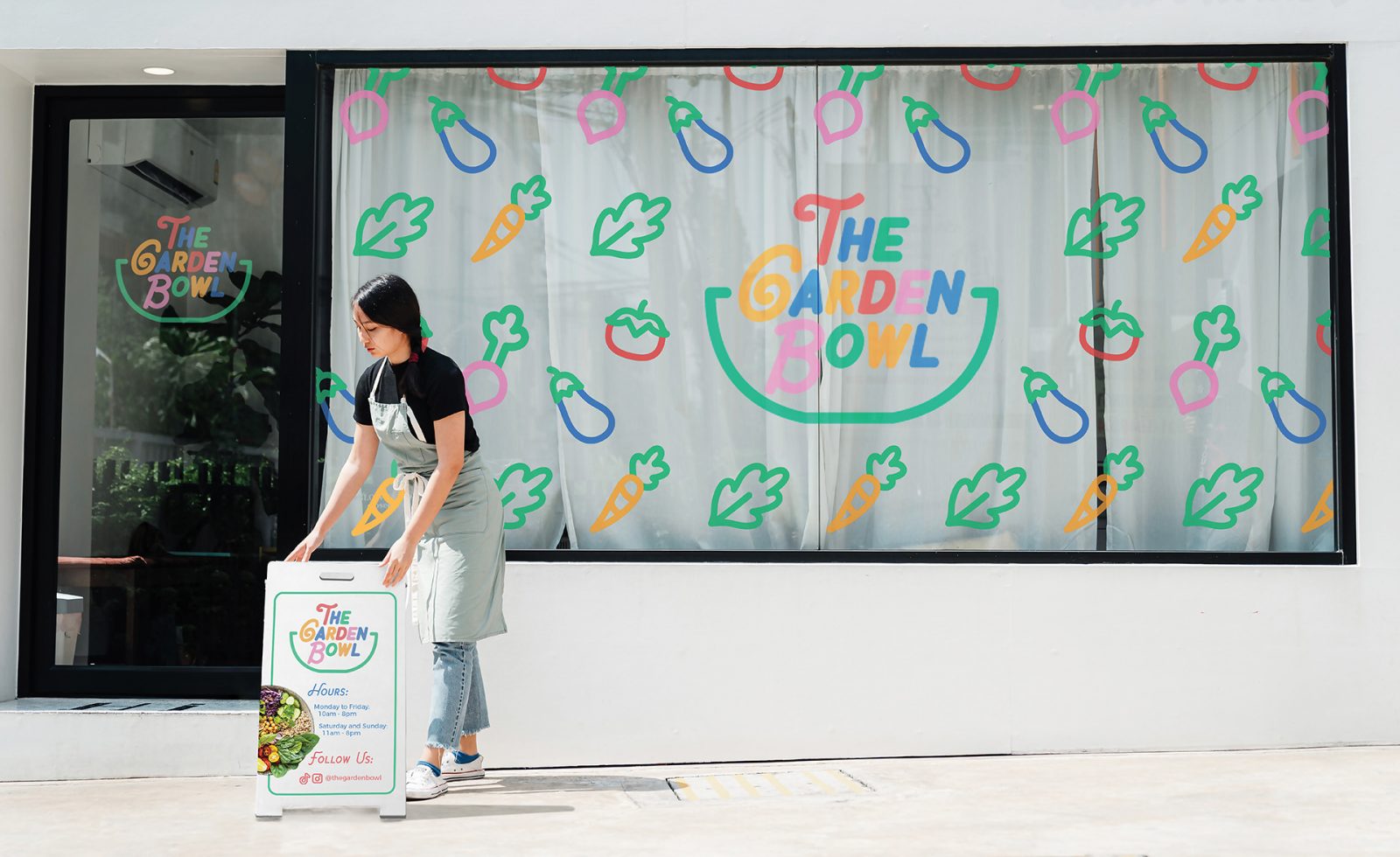 A young woman setting up an open sign outside the front of a colourfully branded restaurant called “The Garden Bowl”. The branding features bright coloured foods such as tomatoes, carrots, and lettuce.